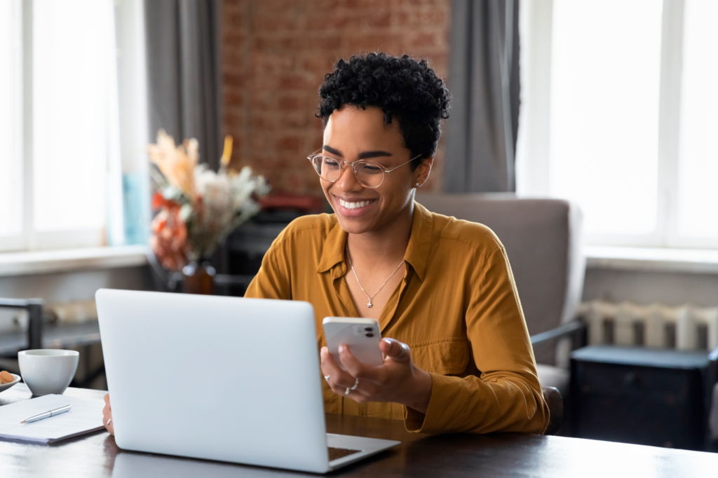 Momentum_African_woman_sit_at_workplace_desk_holds_cellphone_staring_at_laptop__synchronize_data_between_computer_and_gadget_in_office__use_corporate_devices_and_business_application__plan_work__use_organize.png
